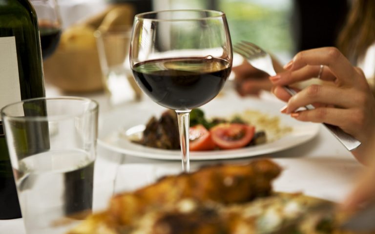 Halal-Catering Berlin mit einem Glas Rotwein und einem Teller mit Gemüse und Fleisch im Hintergrund.